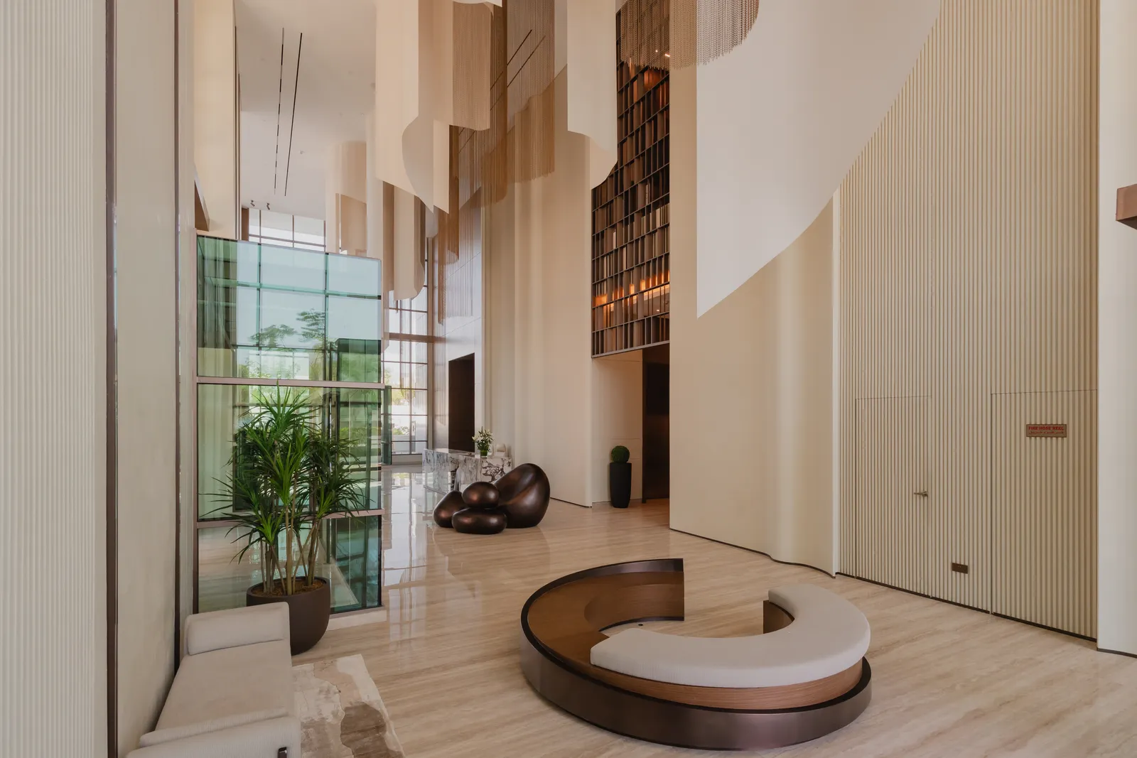The double-height lobby: sheets of pale fabric hanging from the ceiling, a crescent bench in walnut and cream, a cluster of bronze pebble sculptures, a tall bronze lattice screen and a glazed box of planting