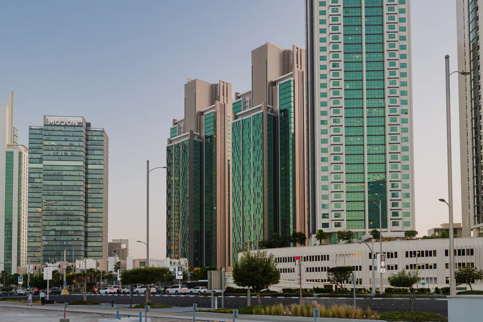 Al Durrah Tower and the matching MAG 5 Residence tower from the street in Marina Square, a taller green tower to their right, an office tower at left, a cream podium building and parked cars in the foreground