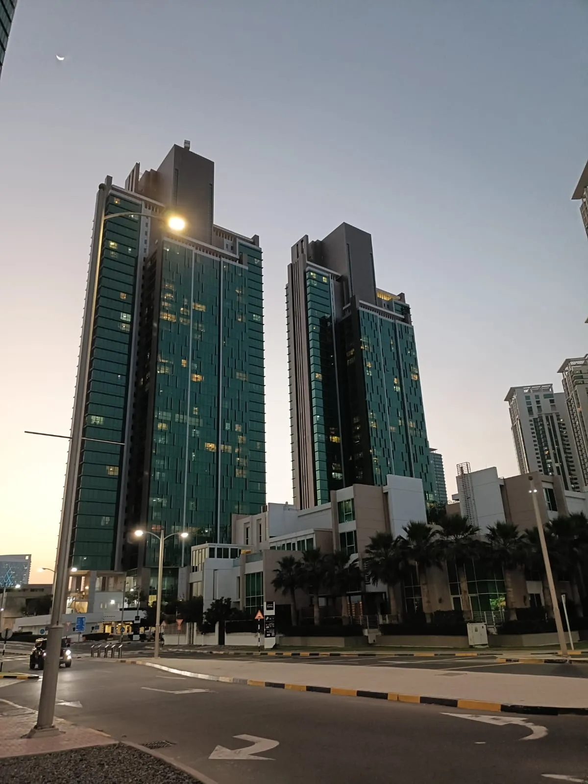 Al Durrah Tower and the matching MAG 5 Residence tower at dusk with lights on in the windows and a crescent moon above, palms and a low building at their base, from a street corner
