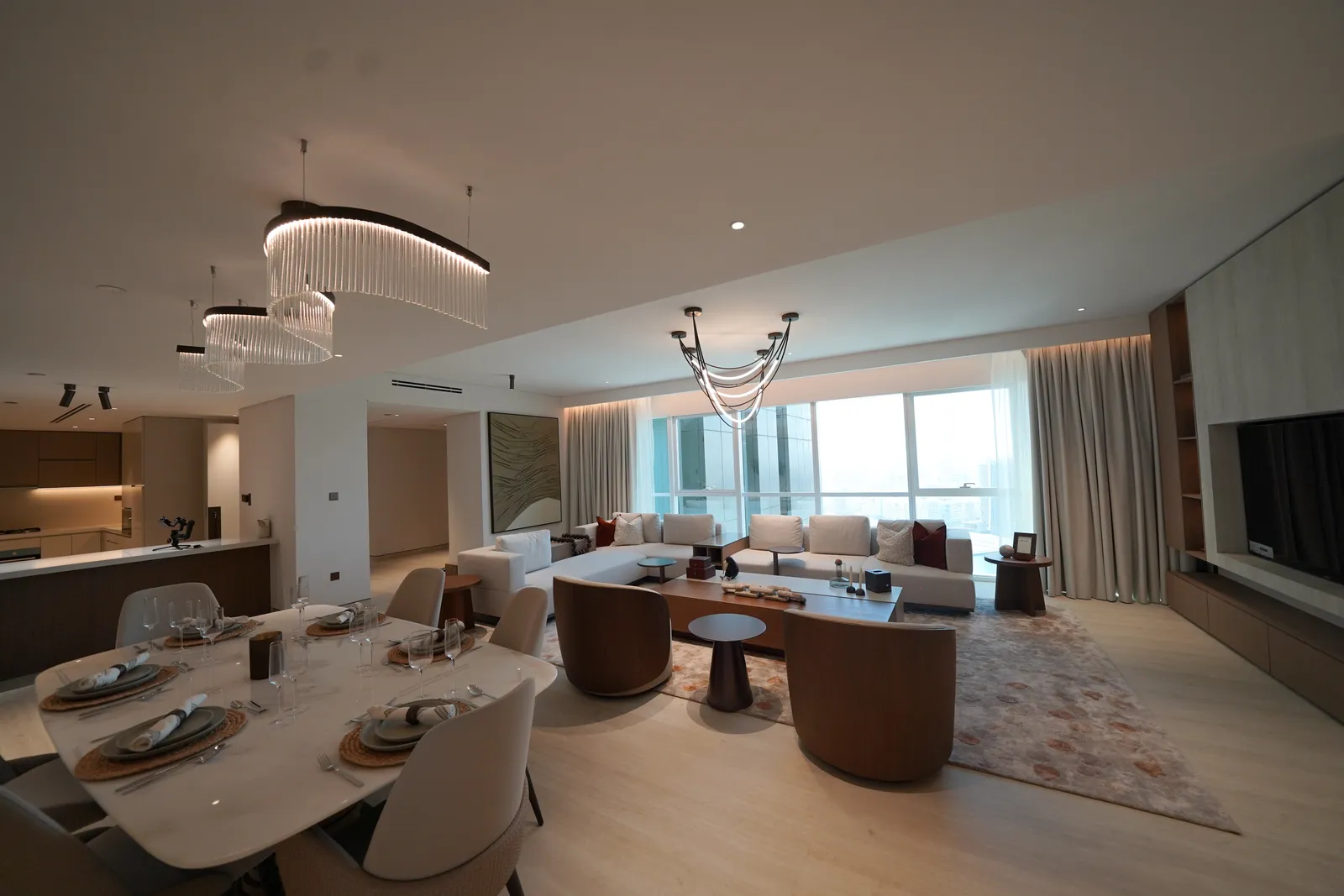 A furnished open-plan living and dining room: a marble table laid for six under a ribbon chandelier, walnut tub chairs, a cream sectional sofa by the window wall, a kitchen counter at left and a pale timber floor