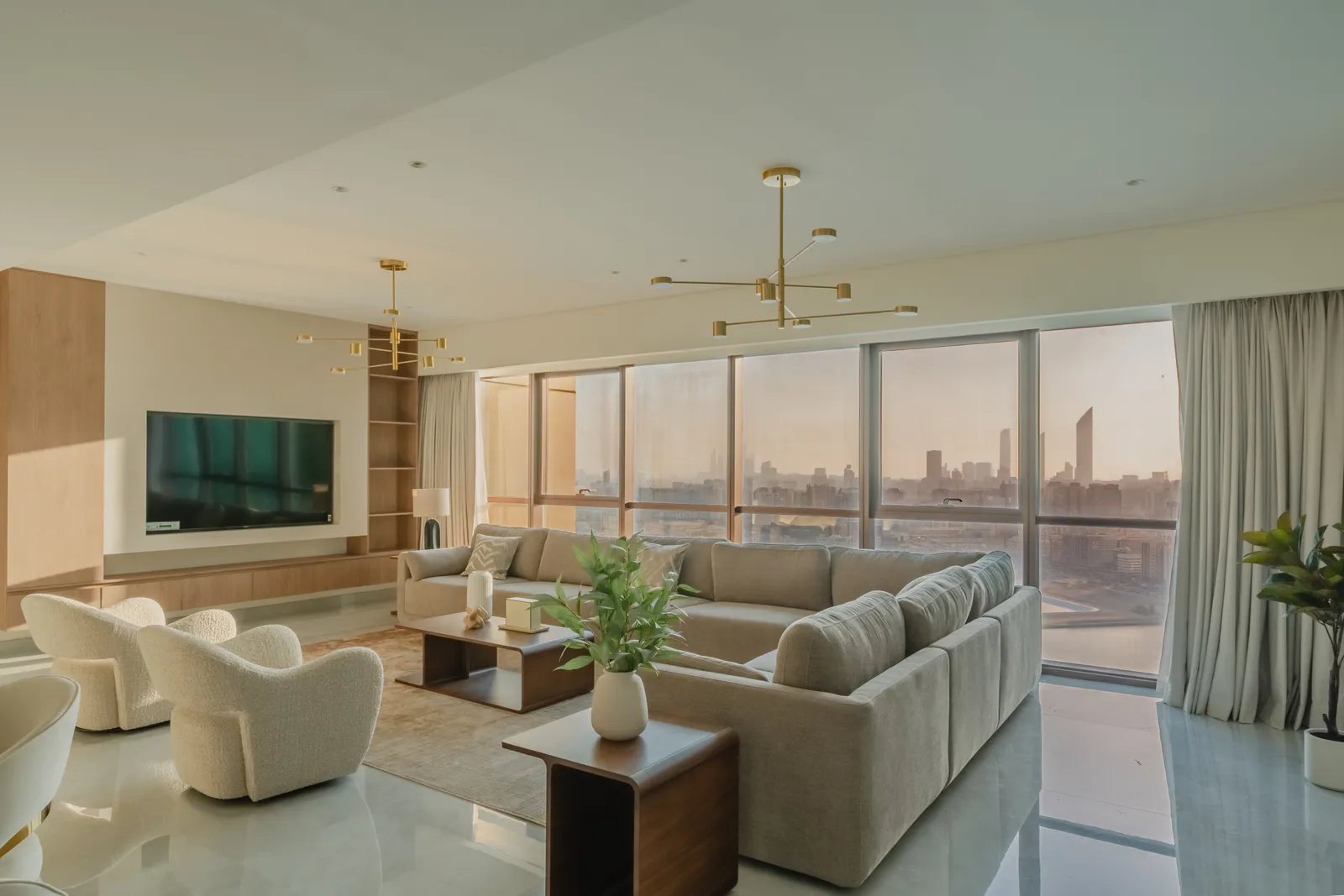 A furnished living room in evening light: a tan sectional sofa, boucle armchairs, brass branched ceiling lights, the skyline through the window wall and a polished floor