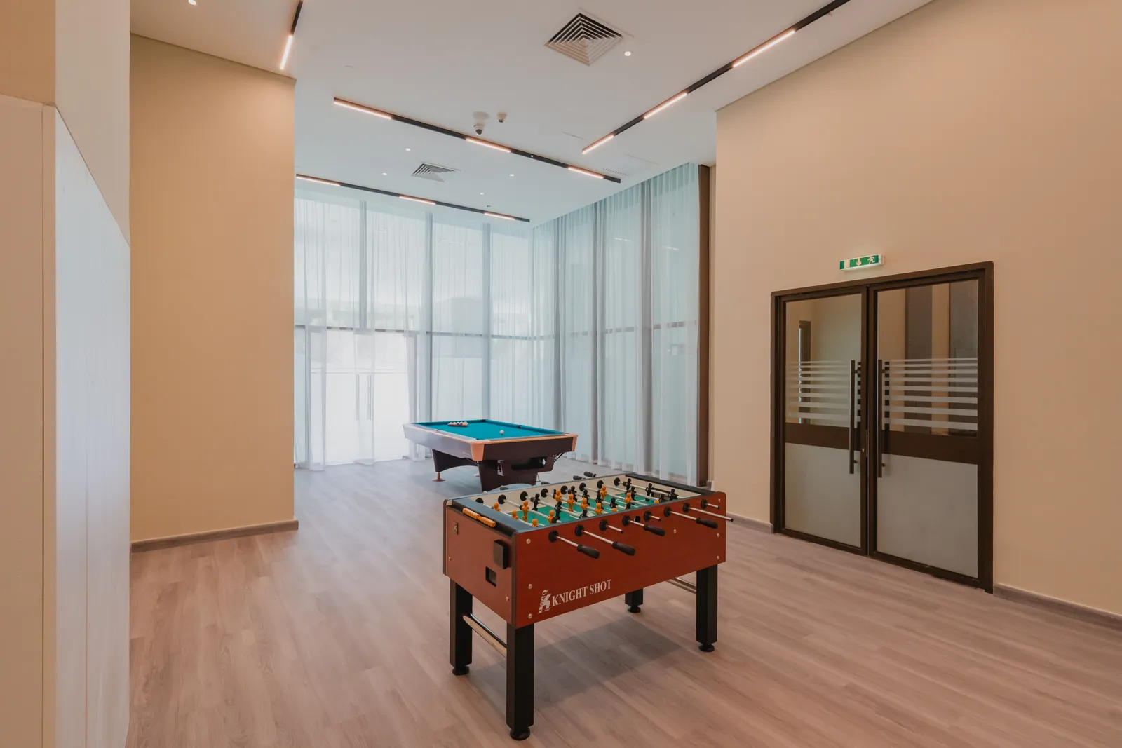 The games room: a table-football table in the foreground, a billiards table by the floor-to-ceiling window with sheer curtains, a glazed double door at right and a light timber floor