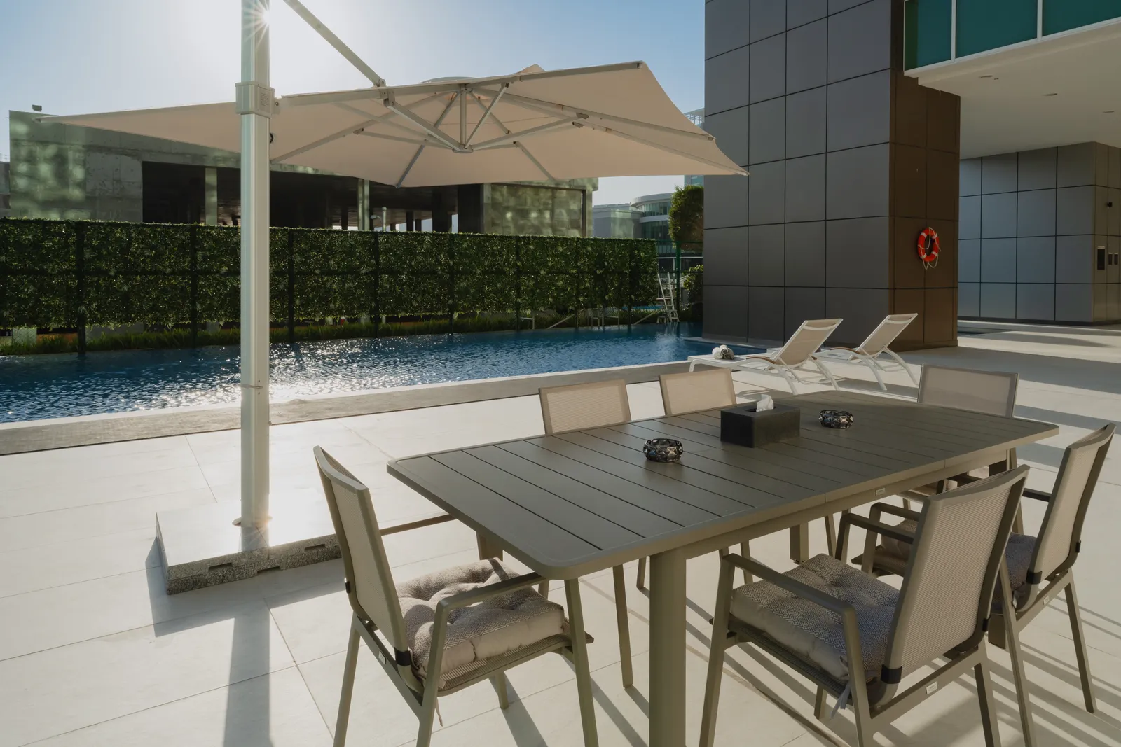An outdoor dining table for six under a cream parasol beside the pool, two loungers by the bronze column with its lifebuoy, the hedge screen beyond