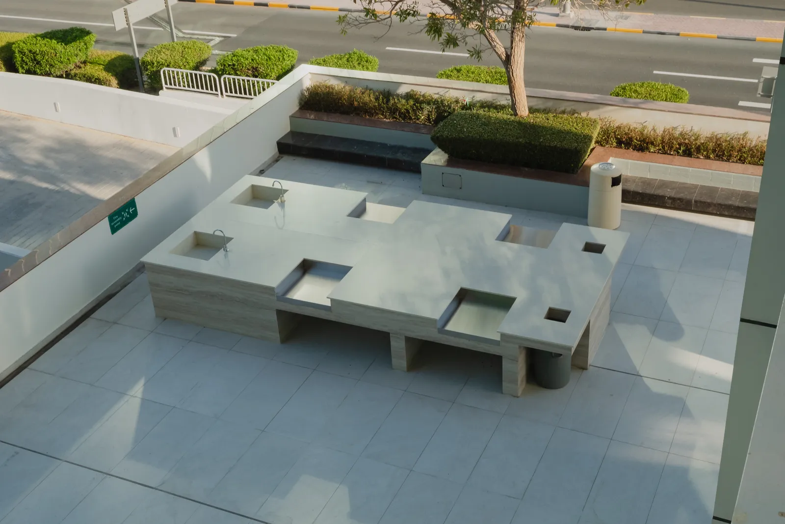 The barbecue terrace from above: a long travertine counter with two sinks and built-in grills, bench seating around it, a planter with a tree and the road beyond the parapet
