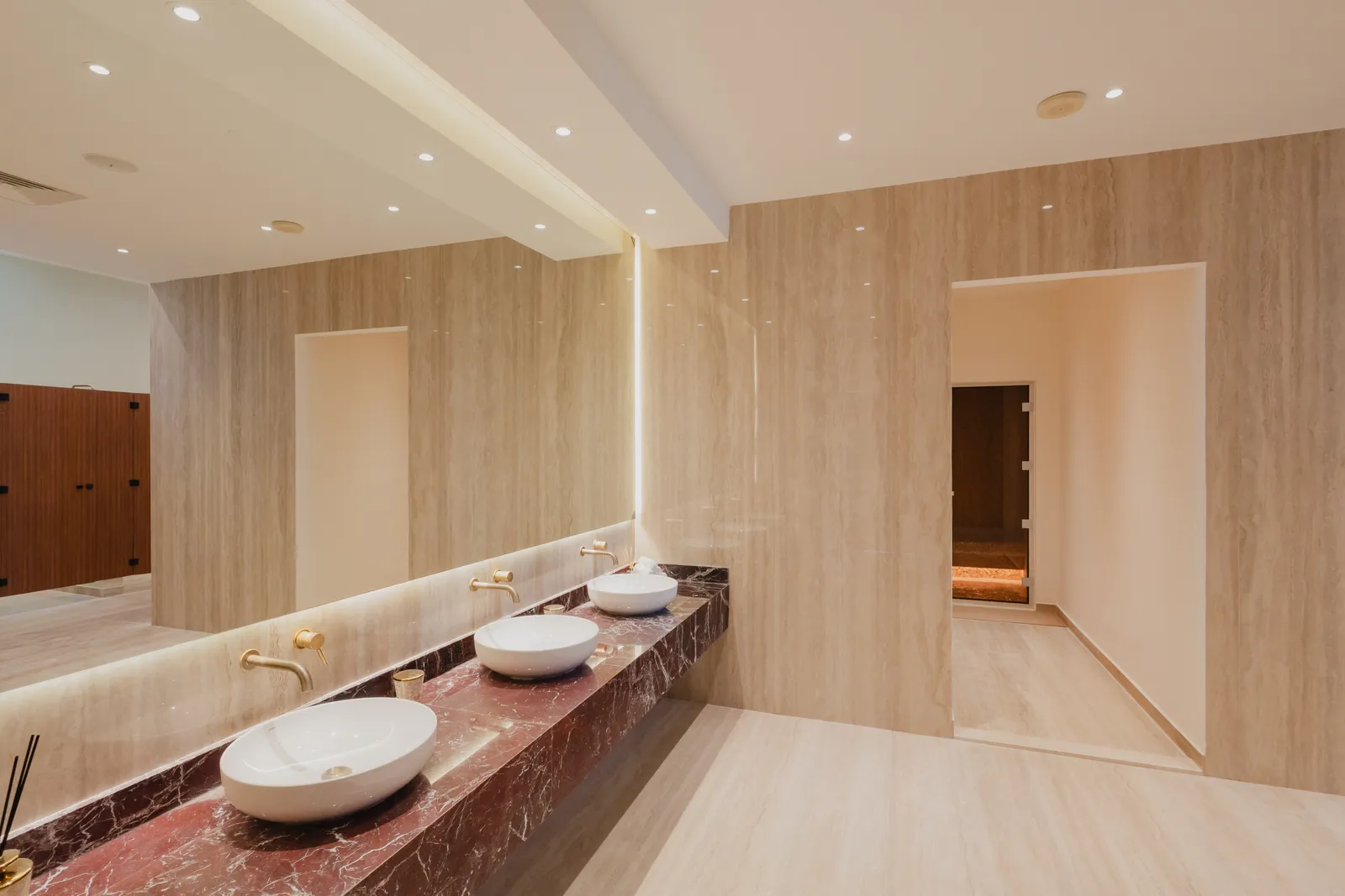 A changing-room vanity: three basins on a red marble counter under a long backlit mirror, travertine walls and a doorway to the sauna