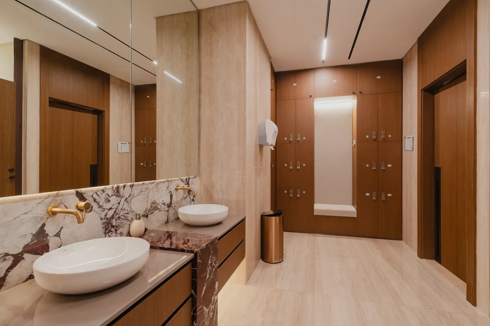 A changing room: a double vanity on a red marble counter, timber lockers with a bench recess, a shower door and a copper bin