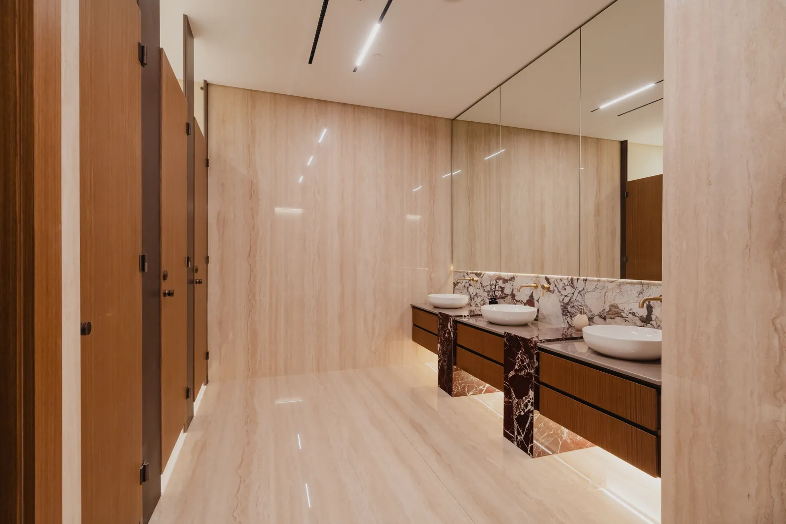 A changing room with timber cubicle doors at left and a mirrored wall above three basins on a veined marble counter