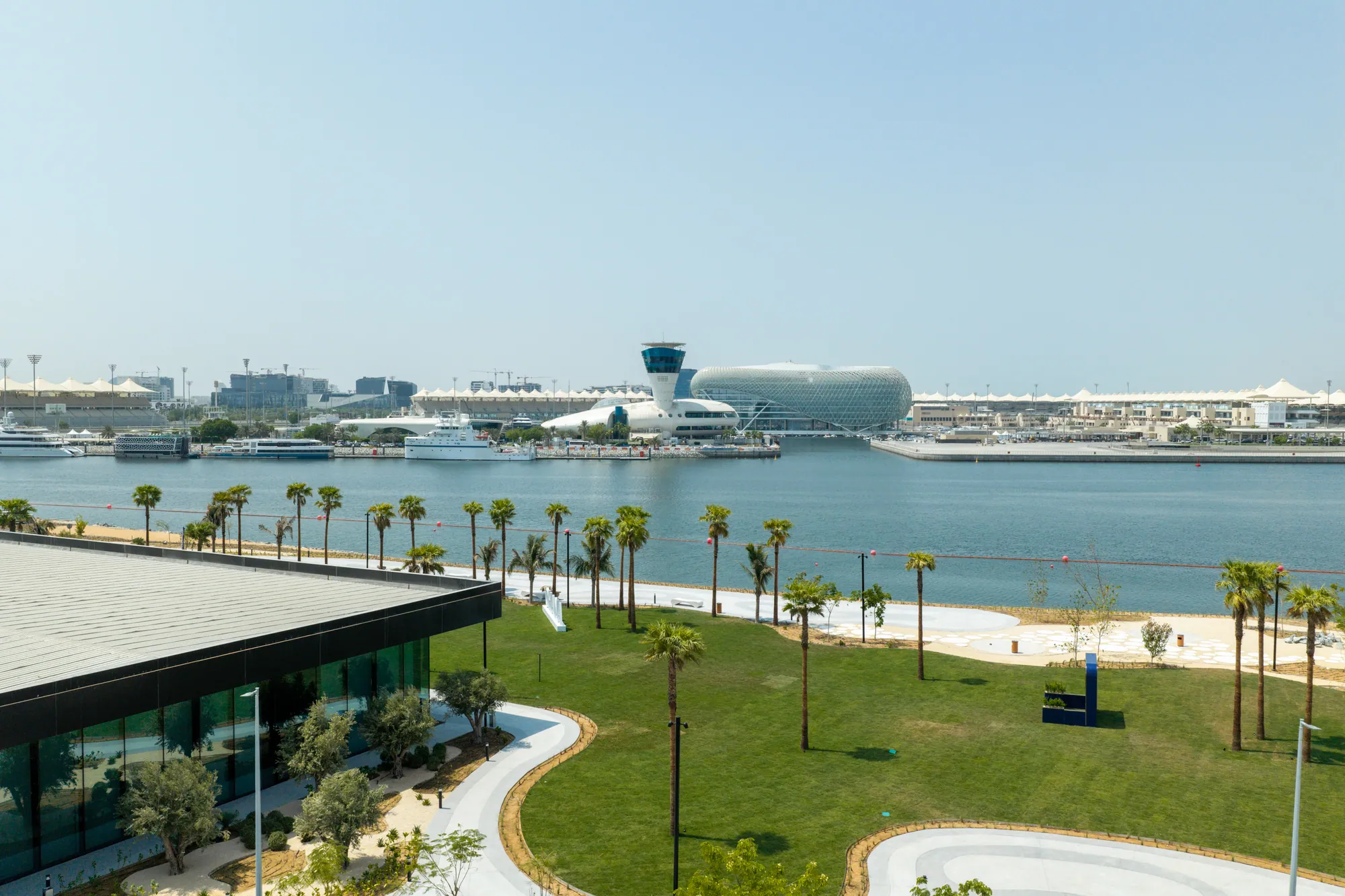 The Yas Canal site from the air on 22 September 2026: the sales centre with a dark roof at the water’s edge, new lawns and paths, palms along the canal, and the Yas Marina Circuit grandstands across the water