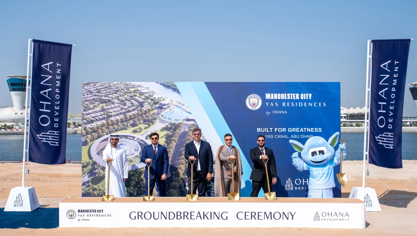 The groundbreaking ceremony on the sand beside the canal: a stage backdrop with the masterplan render and the Manchester City Yas Residences by Ohana lock-up, five people with gold spades and the club mascot, Ohana Development banners either side