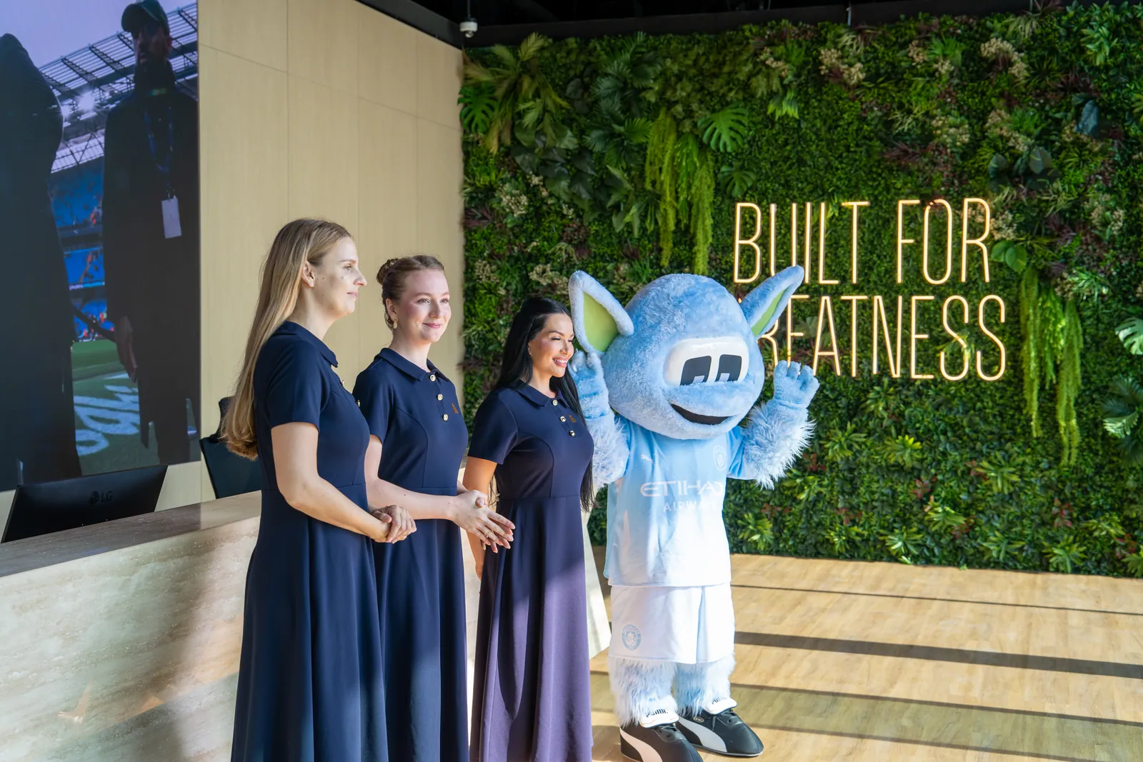Three hostesses in navy dresses beside the club’s light-blue mascot in front of a living green wall lettered Built for Greatness
