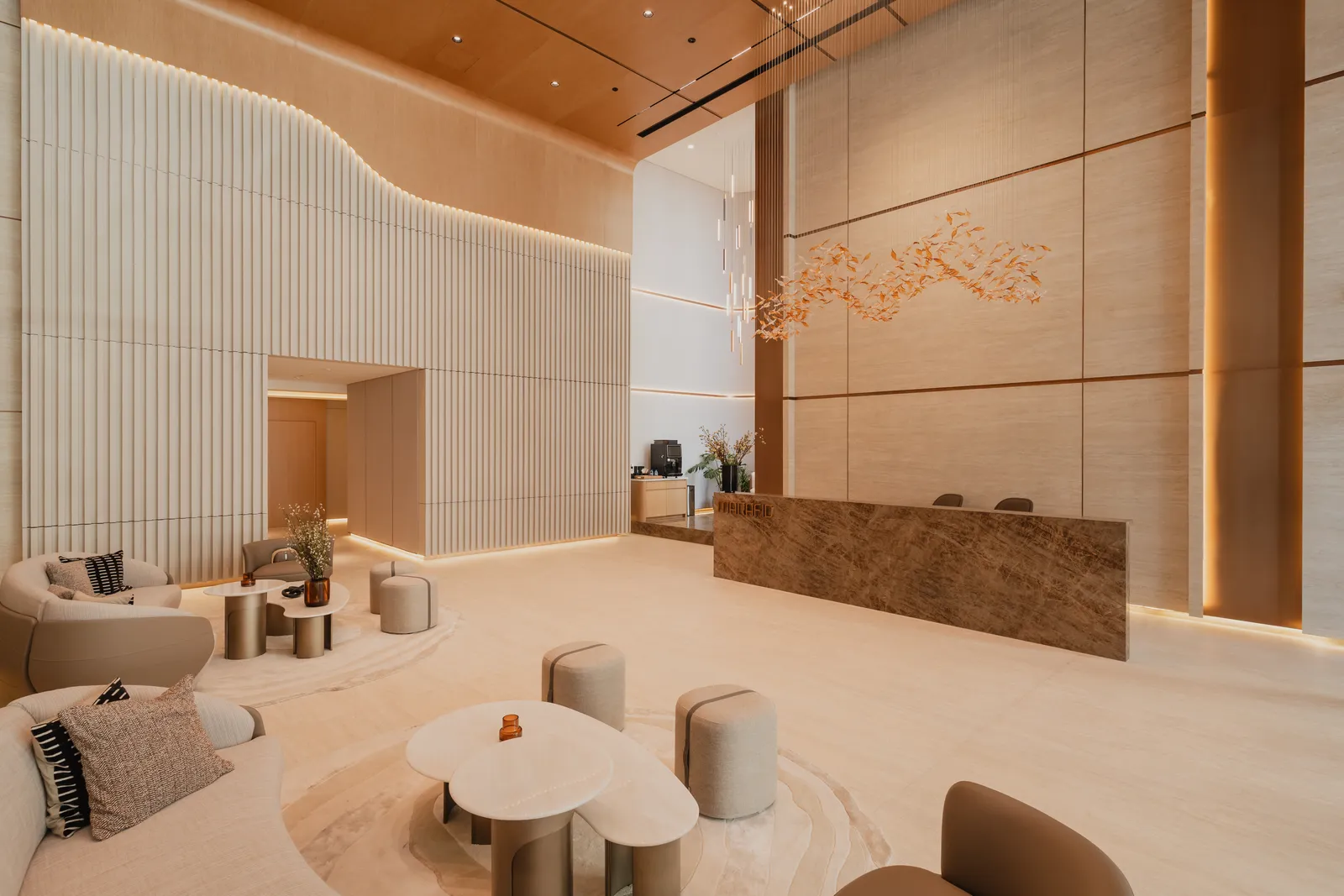The double-height lobby lounge: curved cream sofas and round tables on a rug, a fluted white wall, the marble reception desk and the gold-leaf wall art beyond