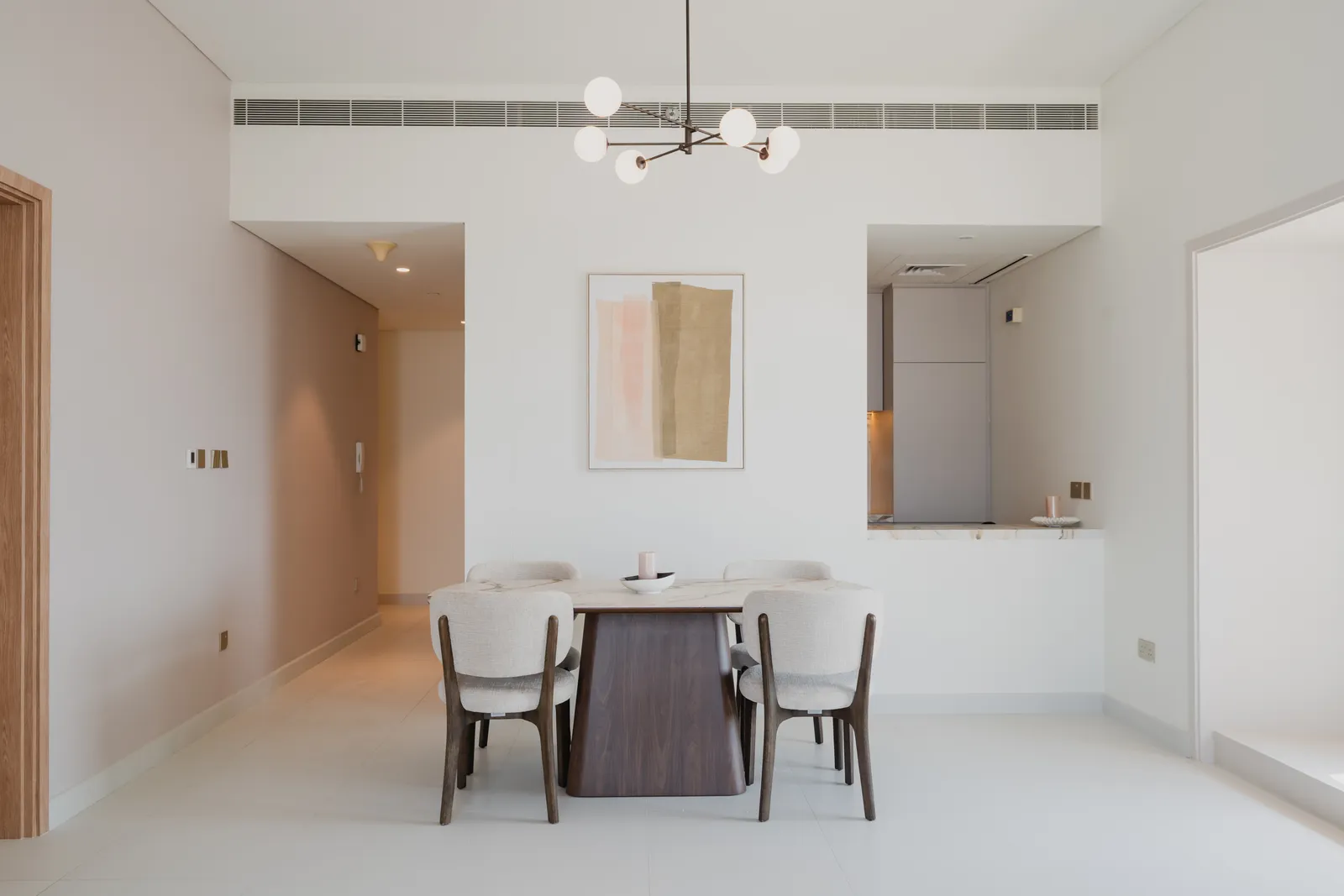 An apartment dining area: a walnut table with four boucle chairs under a branch chandelier, an abstract print on the wall and the kitchen seen through a pass-through