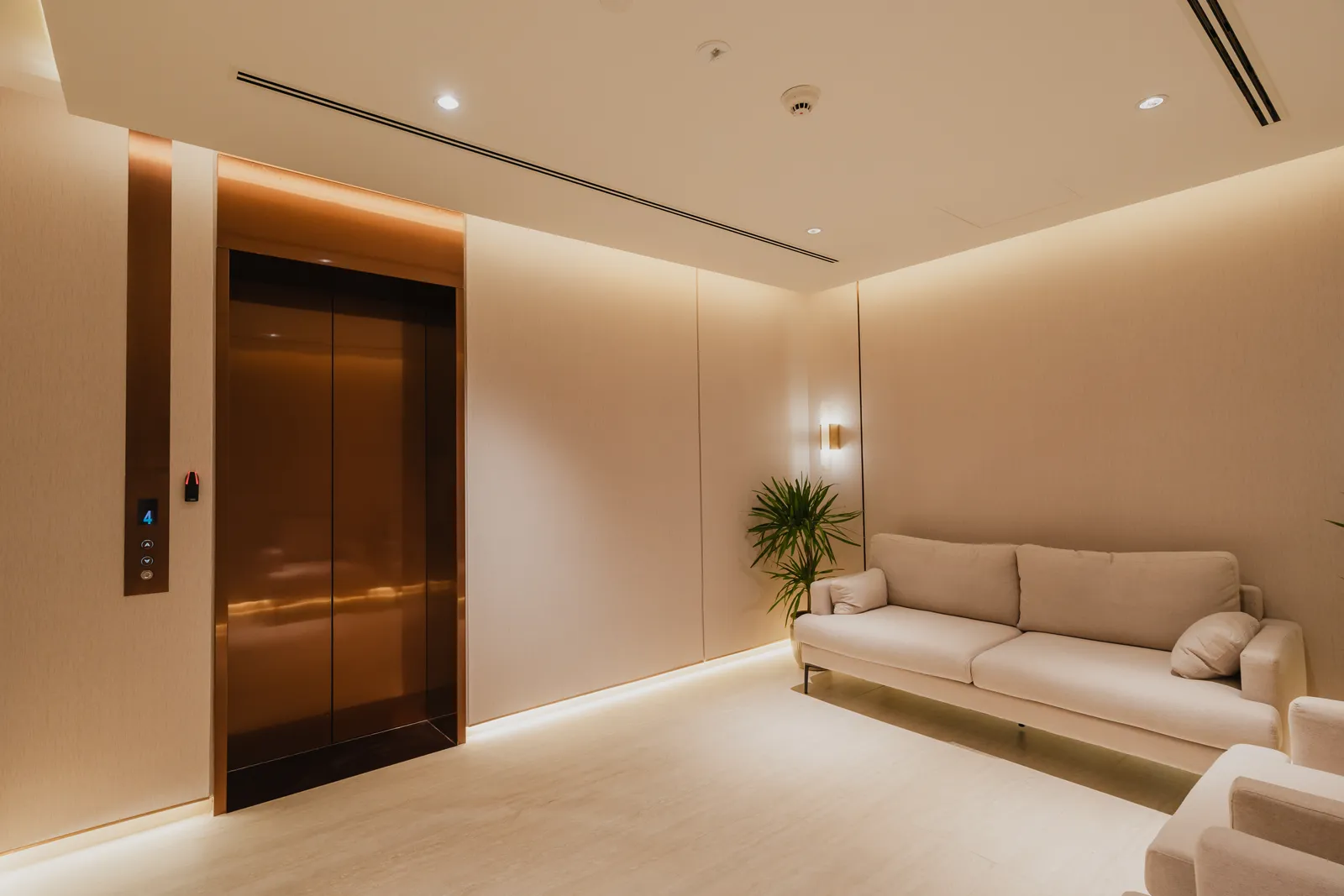 The private lift landing of a three-bedroom apartment: bronze lift doors with the floor indicator reading 4, a cream sofa, a plant and cove lighting