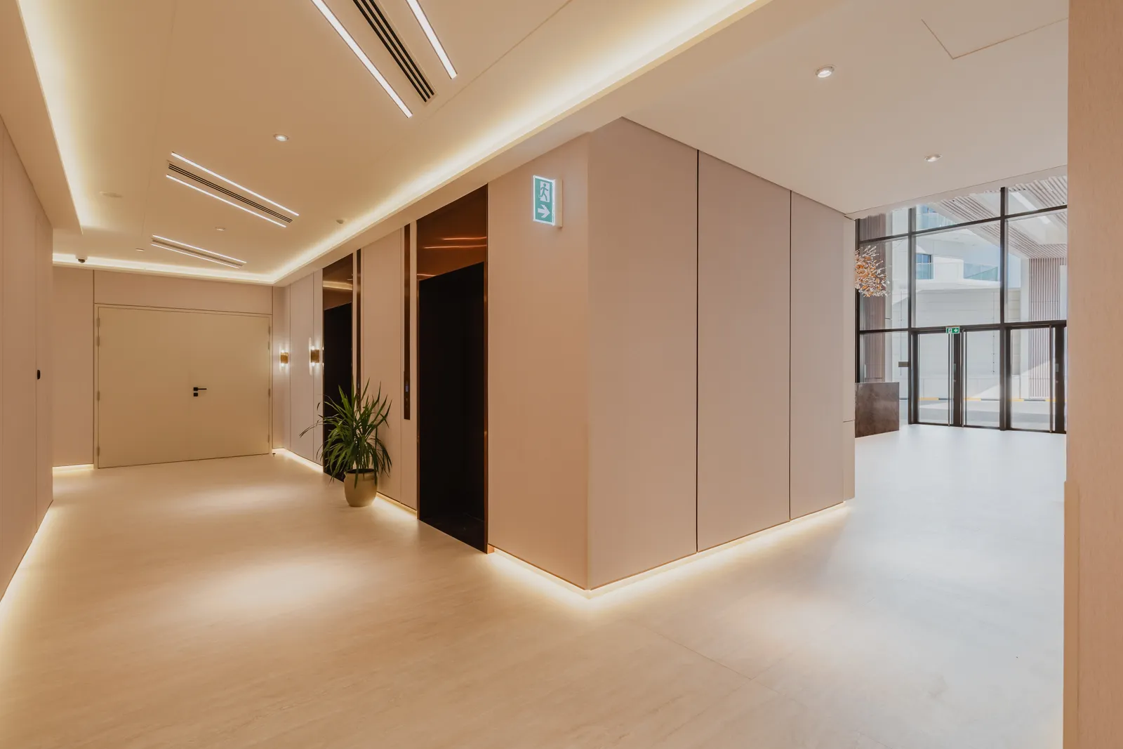 A lift lobby lit from a window at the end, with a door to the side and a bench of cove light along the floor